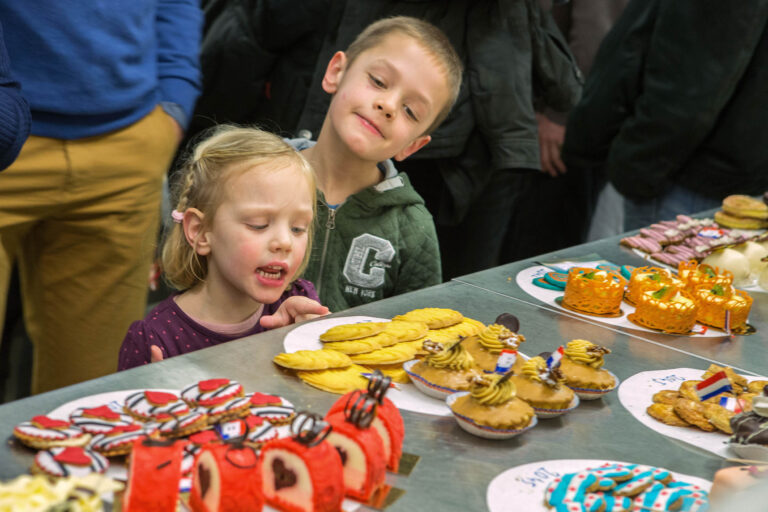 Dit beeld toont twee jonge kinderen die met nieuwsgierigheid kijken naar een uitstalling van kleurrijke gebakjes en koekjes op een tafel. De schotels zijn rijkelijk versierd en uitgestald in verschillende stijlen en kleuren. De kinderen lijken gefascineerd door de lekkernijen voor hen. Om hen heen staan mensen, maar zij zijn gefocust op de zoetigheden. Foto door Jan Buwalda in Groningen.