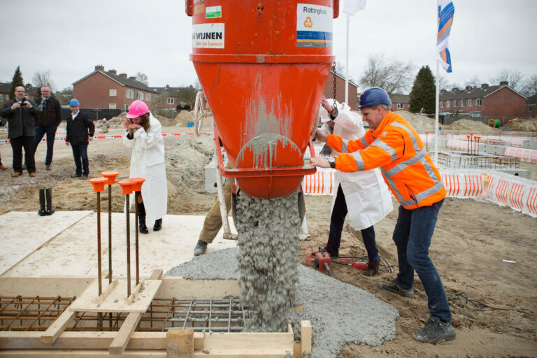 Op de foto zien we een bouwplaats in Groningen waar beton wordt gestort als onderdeel van een constructieproject. Een grote oranje cementemmer leegt zijn inhoud op de voorbereide fundering, terwijl bouwvakkers het proces begeleiden. Een persoon in witte werkkleding en roze veiligheidshelm kijkt toe, terwijl een man in een feloranje veiligheidsjas helpt met storten. Op de achtergrond zijn enkele toeschouwers zichtbaar, evenals omringende woningen. Het geheel suggereert een officiële ceremonie voor de start van de bouw. Foto door Jan Buwalda.