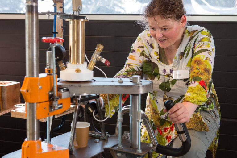 Een vrouw is geconcentreerd bezig met een technische machine. Ze draagt een kleurrijke blouse met bloemenpatroon en bedient een hendel terwijl ze geïnteresseerd kijkt naar het apparaat. Het lijkt een industriële omgeving met veel metalen onderdelen en leidingen. De foto is genomen in een lichte ruimte met daglicht dat door de ramen naar binnen schijnt. Fotograaf: Jan Buwalda, Groningen.