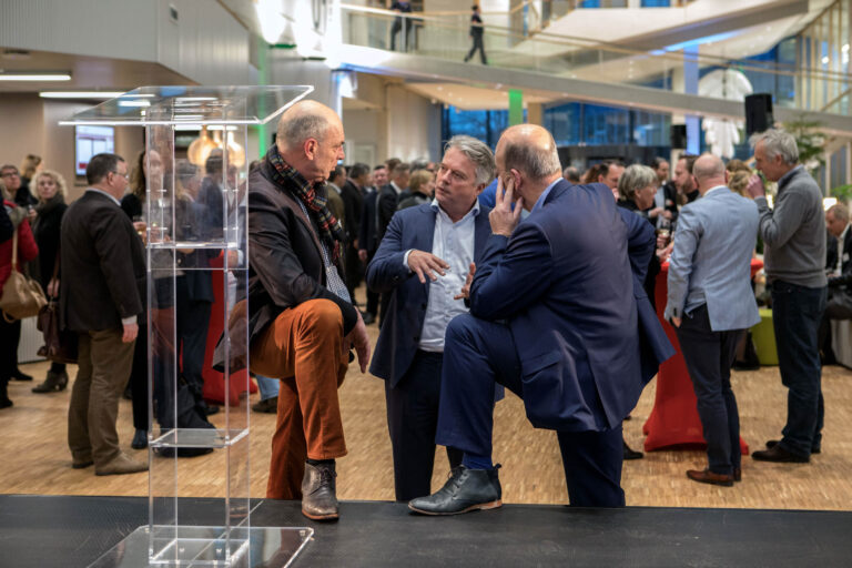 Het beeld toont een druk netwerk-evenement in een moderne ruimte met een mezanine. In het midden zijn drie mannen in pak in een levendige discussie verwikkeld, waarbij de man links een kleurrijke sjaal draagt. Op de achtergrond zijn diverse andere gasten zichtbaar die met elkaar praten in een informele sfeer. De houten vloer en de heldere verlichting dragen bij aan de open en toegankelijke sfeer van de bijeenkomst. Fotograaf: Jan Buwalda.