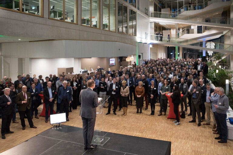 Dit beeld toont een drukbezochte bijeenkomst in een moderne, ruime zaal met meerdere verdiepingen zichtbaar op de achtergrond. De aanwezigen, die netjes gekleed zijn, staan in groepsverband terwijl een spreker op het podium een presentatie of toespraak geeft. De sfeer is professioneel en formeel. De ruimte heeft een open ontwerp met glazen wanden en een houten vloer. Fotograaf: Jan Buwalda, Groningen.