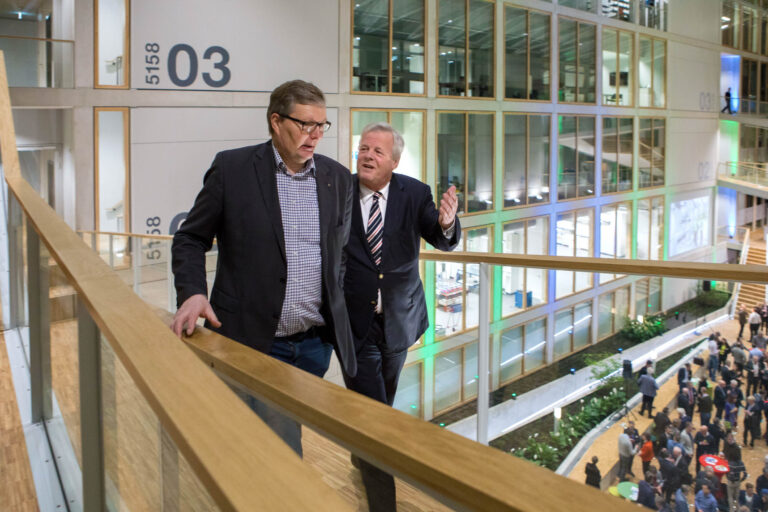 In deze foto, waarvoor Jan Buwalda de fotograaf is, zien we twee mannen in gesprek terwijl ze over een binnenbalkon lopen in een modern kantoorgebouw in Groningen. De ruimte is ruim en licht, met grote ramen en drie verdiepingen genummerd als 03. Op de achtergrond zijn mensen te zien die zich op de begane grond verzamelen. De omgeving lijkt levendig door de open architectuur en het gebruik van glas en hout.