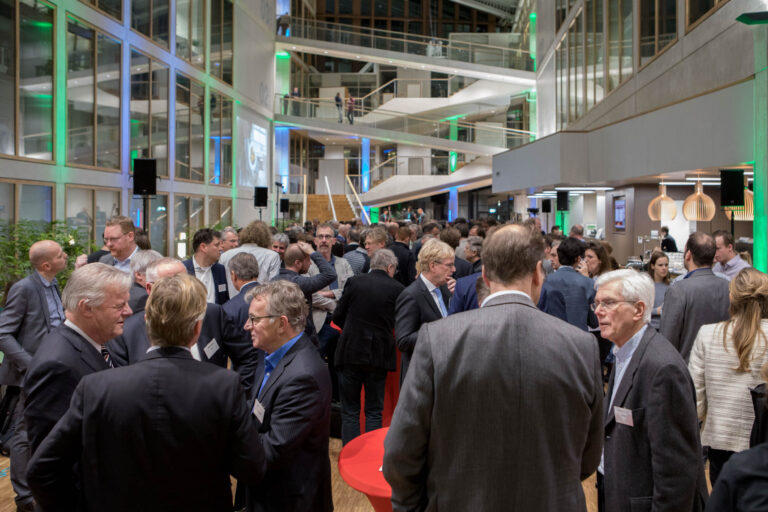 Een grote groep mensen verzameld in een moderne, goed verlichte ontvangsthal, met glazen wanden en een open indeling. Het geroezemoes van gesprekken vult de ruimte terwijl mensen in nette kleding met elkaar in gesprek zijn. De architectuur van het gebouw is opvallend, met meerdere verdiepingen die door middel van trappen verbonden zijn en groene lichtelementen die een sfeervolle ambiance creëren. De omgeving straalt een professionele en zakelijke sfeer uit.Fotograaf: Jan Buwalda, Groningen.