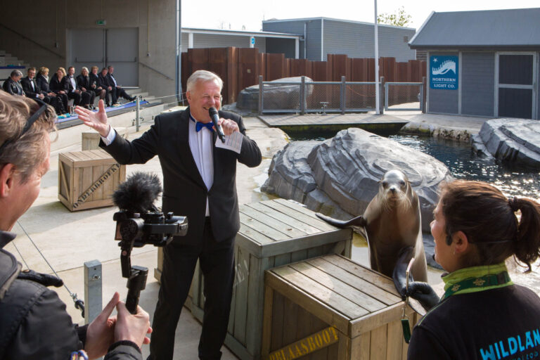 Een evenement in een dierentuin met als centraal thema een zeeleeuwenshow. Op de voorgrond staat een man in een smoking enthousiast te spreken in een microfoon, terwijl een zeeleeuw naast hem op een kist staat, speels kijkend naar een dierverzorger. De omringende mensen zijn toeschouwers die op de achtergrond zitten. De locatie is ingericht met grote rotsen en een zwembad. De foto is genomen door Jan Buwalda in Groningen.