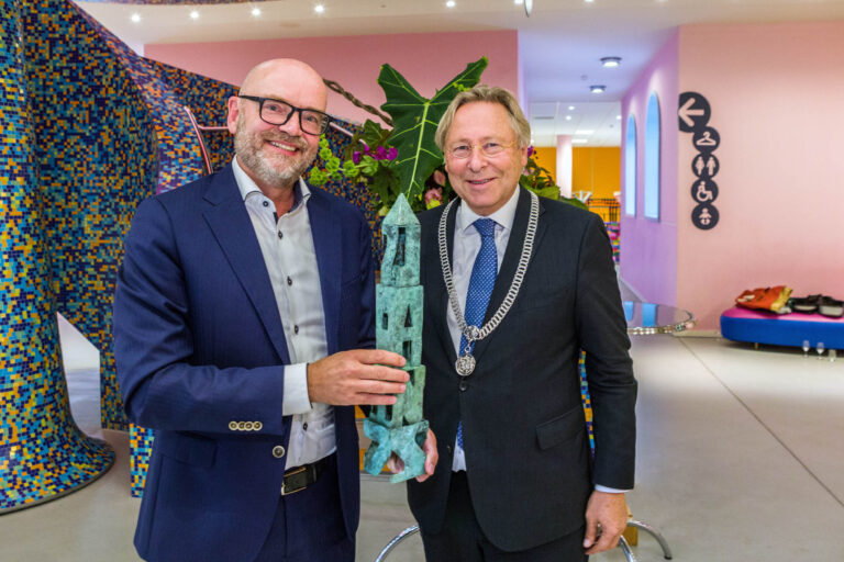 Dit is een foto van twee mannen die samen een sculptuur vasthouden in een kleurrijke en moderne omgeving. De man links draagt een donkerblauw pak en een bril, terwijl de man rechts een ketting draagt die lijkt te behoren bij een officiële functie. Achter hen is een groot kunstwerk of mozaïek zichtbaar met diverse kleuren, en groene planten. De ruimte heeft zachtroze muren en een speelse inrichting. Fotograaf: Jan Buwalda, Groningen.
