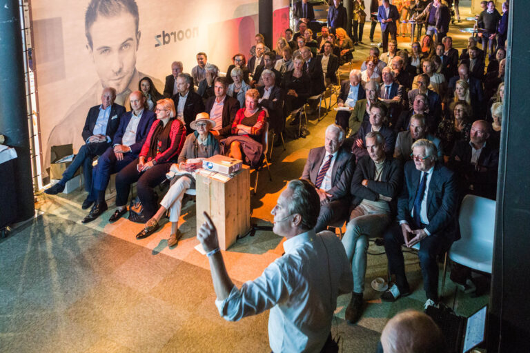 Deze foto, gemaakt door Jan Buwalda in Groningen, toont een groep mensen die aandachtig luisteren naar een presentatie. De spreker staat vooraan met een microfoon, terwijl het publiek geduldig op hun stoelen zit. De setting lijkt professioneel en daar is een grote poster op de achtergrond zichtbaar. De sfeer is geconcentreerd en de verlichting benadrukt de spreker en de voorste rijen.