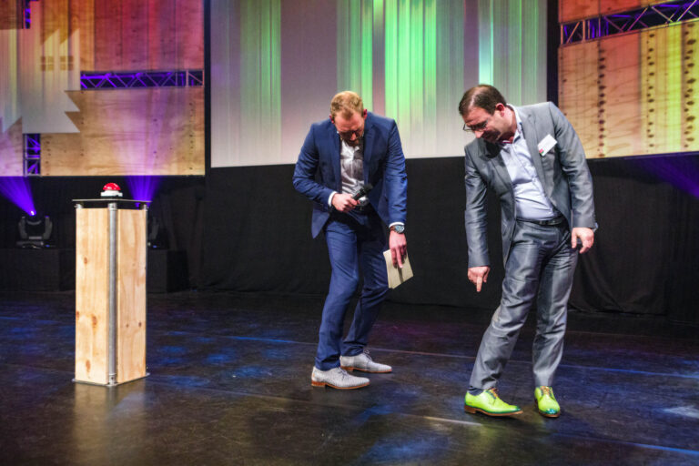 Twee mannen in pakken staan op een podium tijdens een evenement. De man links houdt een microfoon vast en kijkt naar de grond, terwijl de man rechts met een glimlach naar zijn opvallende groene schoenen wijst. Het podium is omgeven door kleurrijke lichteffecten en een houten standaard met een rode knop erop. Foto door Jan Buwalda, Groningen.
