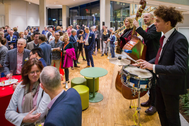 In deze levendige foto, genomen door Jan Buwalda in Groningen, is een drukke receptie te zien in een moderne binnenruimte. De vloer van licht hout en de glazen wanden creëren een open sfeer. Een live band speelt op de voorgrond met een drummer, contrabassist en gitarist, die voor een feestelijke muzikale ambiance zorgen. De gasten lijken te genieten van elkaars gezelschap, met velen zij-aan-zij in gesprek, terwijl ze een glas vasthouden. De rode en groene accenten in de meubels voegen kleur toe aan de scène.