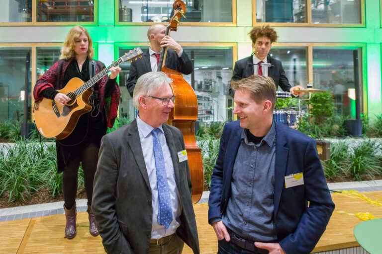 Dit beeld, vastgelegd door fotograaf Jan Buwalda in Groningen, toont een levendig tafereel. Op de voorgrond zijn twee heren in gesprek, beiden gekleed in een net pak, uitstralend een vriendelijke en informele sfeer. Op de achtergrond speelt een ensemble van drie muzikanten: een gitariste, een bassist, en een drummer. De muzikanten zijn geconcentreerd op hun optreden, en de kleurrijke verlichting en de groene planten creëren een aangename ambiance. Het geheel straalt een gevoel van gezelligheid en levendigheid uit tijdens een evenement.