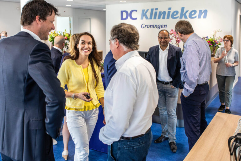 Dit beeld toont een groep mensen die een informele bijeenkomst bijwoont in de ontvangstruimte van DC Klinieken in Groningen. De deelnemers lijken ontspannen en in gesprek met elkaar in een lichte en moderne omgeving. Op de achtergrond is er een bord met de naam van de kliniek zichtbaar. Het lijkt een professionele setting te zijn met een vriendelijke sfeer. Deze foto is genomen door Jan Buwalda.