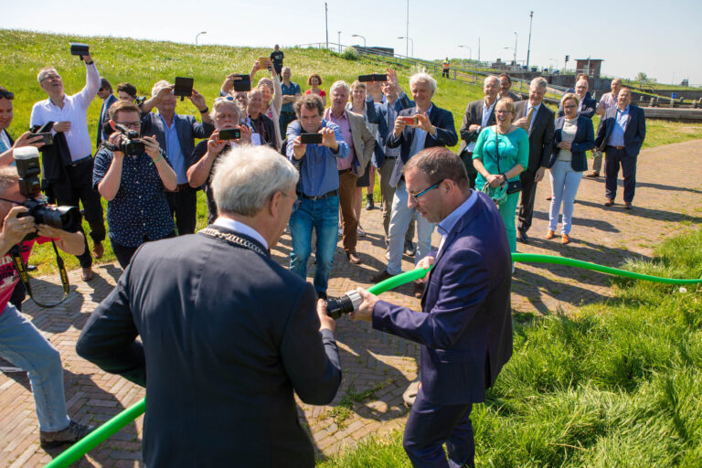 Een groep mensen is verzameld op een zonnige dag om een belangrijk moment vast te leggen. Op de voorgrond zijn twee mannen te zien die een groen lint hanteren. Om hen heen staan mensen met camera's en smartphones om het tafereel te fotograferen. De achtergrond toont een groene dijk en een heldere blauwe lucht. De sfeer is feestelijk en offcieel. Foto door Jan Buwalda, Groningen.