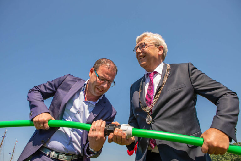 Ik kan de personen op de foto niet herkennen, maar hier is een beschrijving van de afbeelding:Op een zonnige dag zijn twee mannen in formele pakken bezig met een groene slang of buis. Ze lijken samen te werken aan het bevestigen ervan. De lucht is helder blauw, wat zorgt voor een frisse en energieke sfeer. Het is een moment van samenwerking en collegialiteit. Deze foto is genomen door fotograaf Jan Buwalda in Groningen.