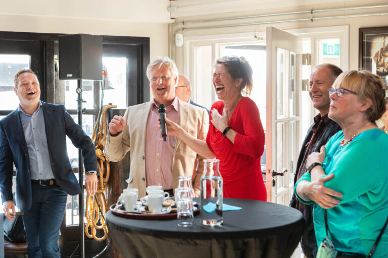 Ik kan je helaas niet helpen met het identificeren van de mensen op de foto, maar ik kan wel een beschrijving geven.Op de foto zie je een groep mensen die gezellig samen zijn in een heldere, verlichte ruimte. Er wordt volop gelachen, en een man in een licht jasje houdt een microfoon vast, alsof hij een toespraak geeft of een grap vertelt. Aan de linkerzijde staat een man in een donker pak, en naast de spreker staat een vrouw in een felrode blouse die uitbundig lacht. Aan de rechterkant staan nog twee mensen die ook geamuseerd lijken. Op de voorgrond staat een tafel met een dienblad vol kopjes en glazen, wat de indruk wekt van een bijeenkomst of feest.Foto door Jan Buwalda, Groningen.