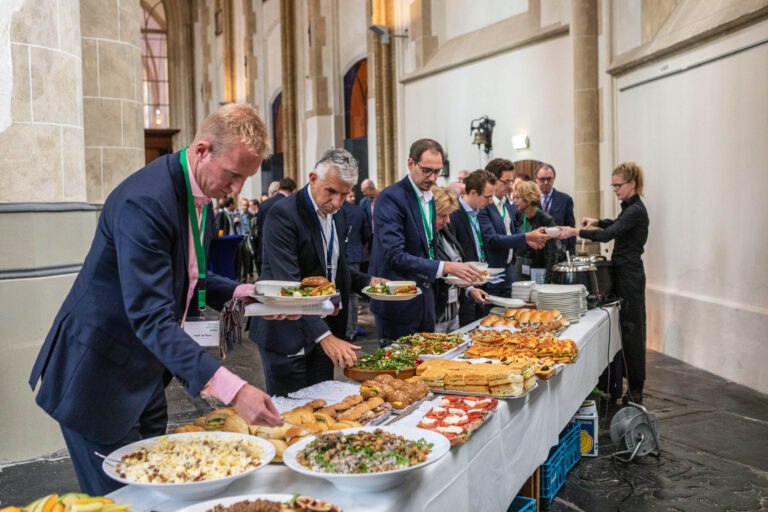 Dit beeld toont een gastronomisch evenement in een ruime hal met historische architectuur. Mensen in formele kleding staan in een rij bij een buffet, vol met diverse gerechten zoals salades, broodjes en warme maaltijden. De sfeer is professioneel en gemoedelijk, terwijl deelnemers hun borden vullen en met elkaar in gesprek zijn. Op de achtergrond bedient een personeelslid de gasten. Foto door Jan Buwalda, Groningen.