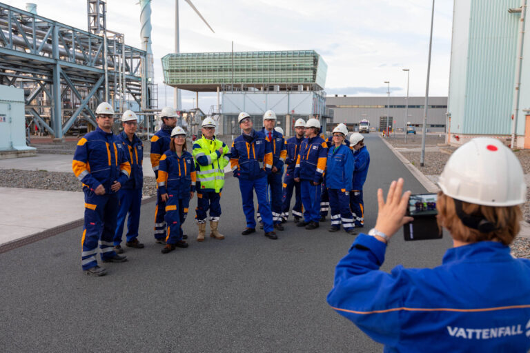 Dit is een foto van een groep werknemers in blauwe en oranje veiligheidskleding en helmen, genomen op een industrieterrein. Op de achtergrond staan industriële gebouwen en een windturbine. Een persoon maakt een foto van de groep met een tablet. Deze foto is gemaakt door Jan Buwalda in Groningen.