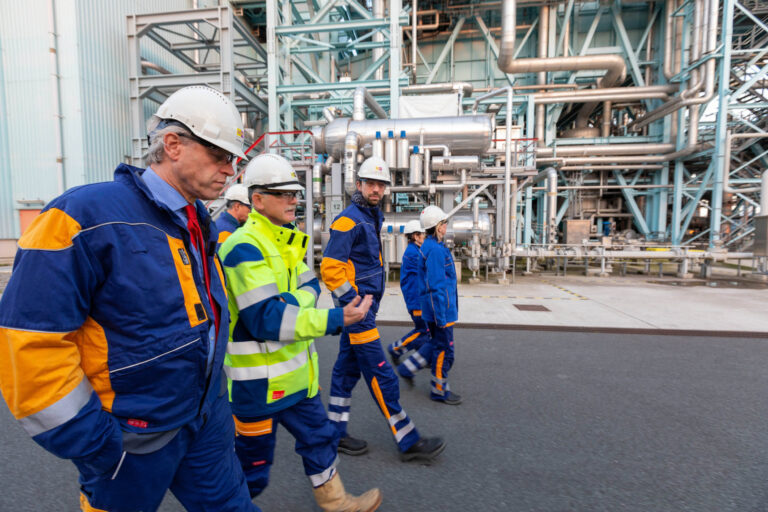 Ik zie een groep mensen in veiligheidshelmen en werkkleding die langs een industriële installatie lopen. De achtergrond toont een complex netwerk van pijpleidingen en structuren, typisch voor een fabriek. Dit is waarschijnlijk een rondleiding of inspectie. Foto door Jan Buwalda, genomen in Groningen.