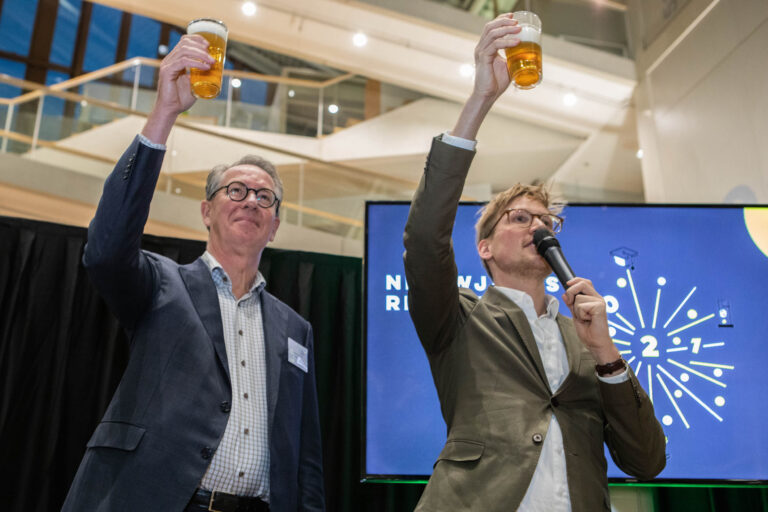 In de foto genomen door Jan Buwalda in Groningen, houden twee heren een glas bier omhoog, alsof ze proosten. De man rechts houdt bovendien een microfoon vast. De sfeer oogt feestelijk en er is een scherm op de achtergrond met feestelijke symbolen. De locatie lijkt een modern interieur met veel lichtinval.