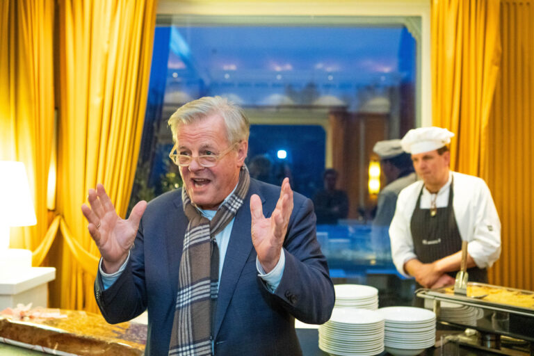 Dit is een levendige foto van een man in een chique ruimte met gele gordijnen. Hij draagt een blauw pak en een geruite sjaal, en lijkt enthousiast aan het spreken te zijn, met zijn handen in gebaren. Op de achtergrond staat een kok in uniform naast een tafel gedekt met witte borden. De sfeer is informeel en gemoedelijk. Fotografie door Jan Buwalda uit Groningen.