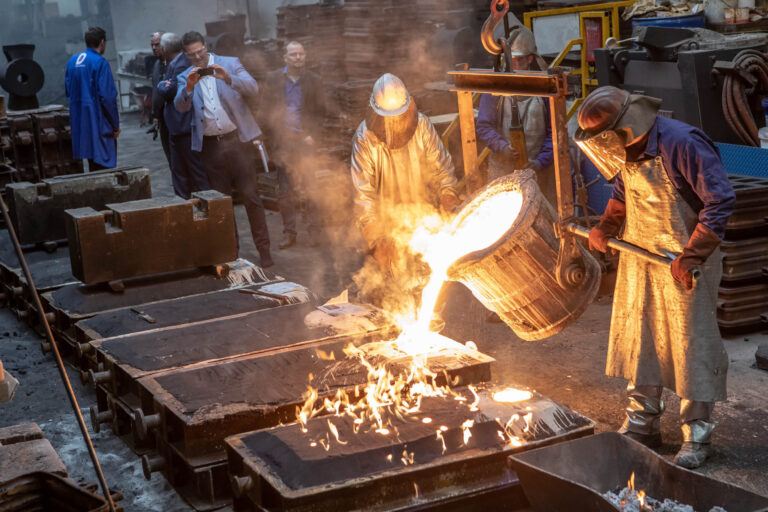 Dit indrukwekkende beeld toont een smelterij waar werknemers in beschermende kleding gloeiend heet gesmolten metaal in vormen gieten. De vuurzee en de vlammen geven een krachtige indruk van de hitte en energie in deze industriële omgeving. Op de achtergrond staan enkele toeschouwers, mogelijk belangstellenden of collega’s, die het proces nauwkeurig volgen. De foto is genomen door Jan Buwalda in Groningen.