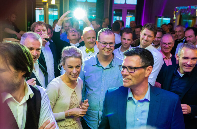 Deze foto, gemaakt door Jan Buwalda in Groningen, toont een groep mensen die zich amuseren op een feestje of bijeenkomst. De sfeer is levendig en vrolijk, met veel lachende gezichten. Op de achtergrond zie je een flits van een camera, wat de feestelijke ambiance benadrukt. De aanwezigen zijn stijlvol gekleed, wat suggereert dat het een speciale gelegenheid betreft.