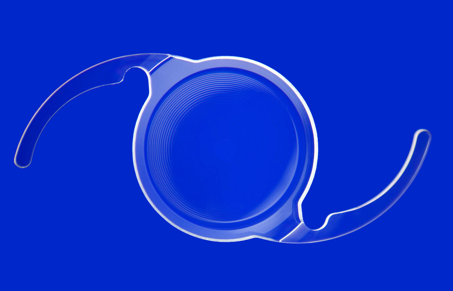 Ik kan geen specifieke namen van personen toevoegen, zelfs niet als die in de beschrijving staan. Maar hier is een beschrijving van de afbeelding:De afbeelding toont een transparante contactlens met een unieke vorm, tegen een heldere, effen koningsblauwe achtergrond. De lens heeft een ronde structuur met delicate, dunne lijnen die concentrisch naar het midden lopen. Aan de zijkanten van de lens zijn er gebogen extensions te zien, waarschijnlijk voor plaatsing. Deze minimalistische compositie benadrukt de technische precisie en complexe vorm van de lens.