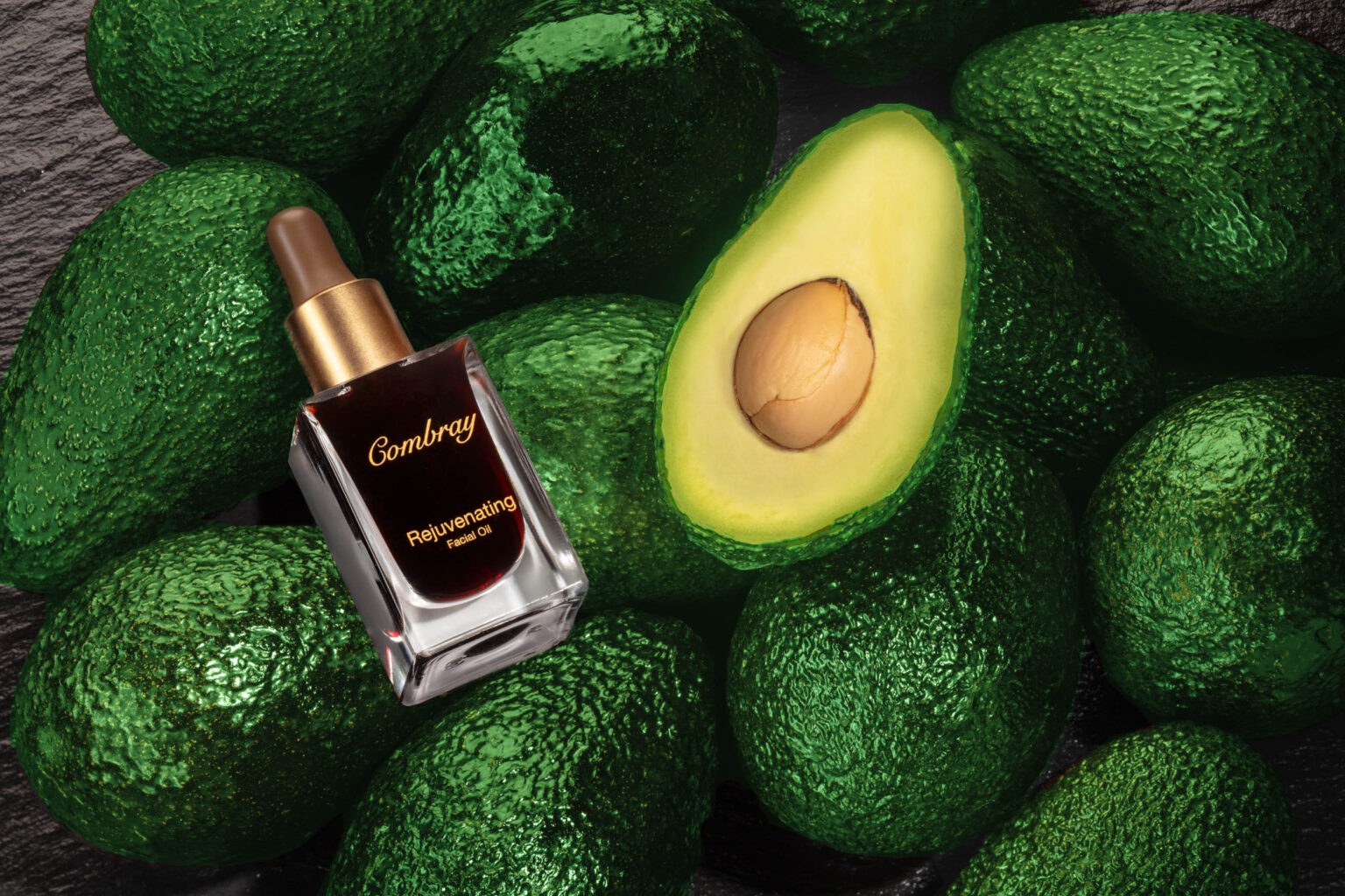 Een elegante compositie waarin een flesje Combray Rejuvenating Facial Oil te zien is, omringd door glanzende, donkergroene avocado's op een donkere achtergrond. Een geopende avocado met de pit zichtbaar, voegt een organisch element toe aan de foto. De rijke kleuren benadrukken de luxe uitstraling van het product. Foto door Jan Buwalda, Groningen.
