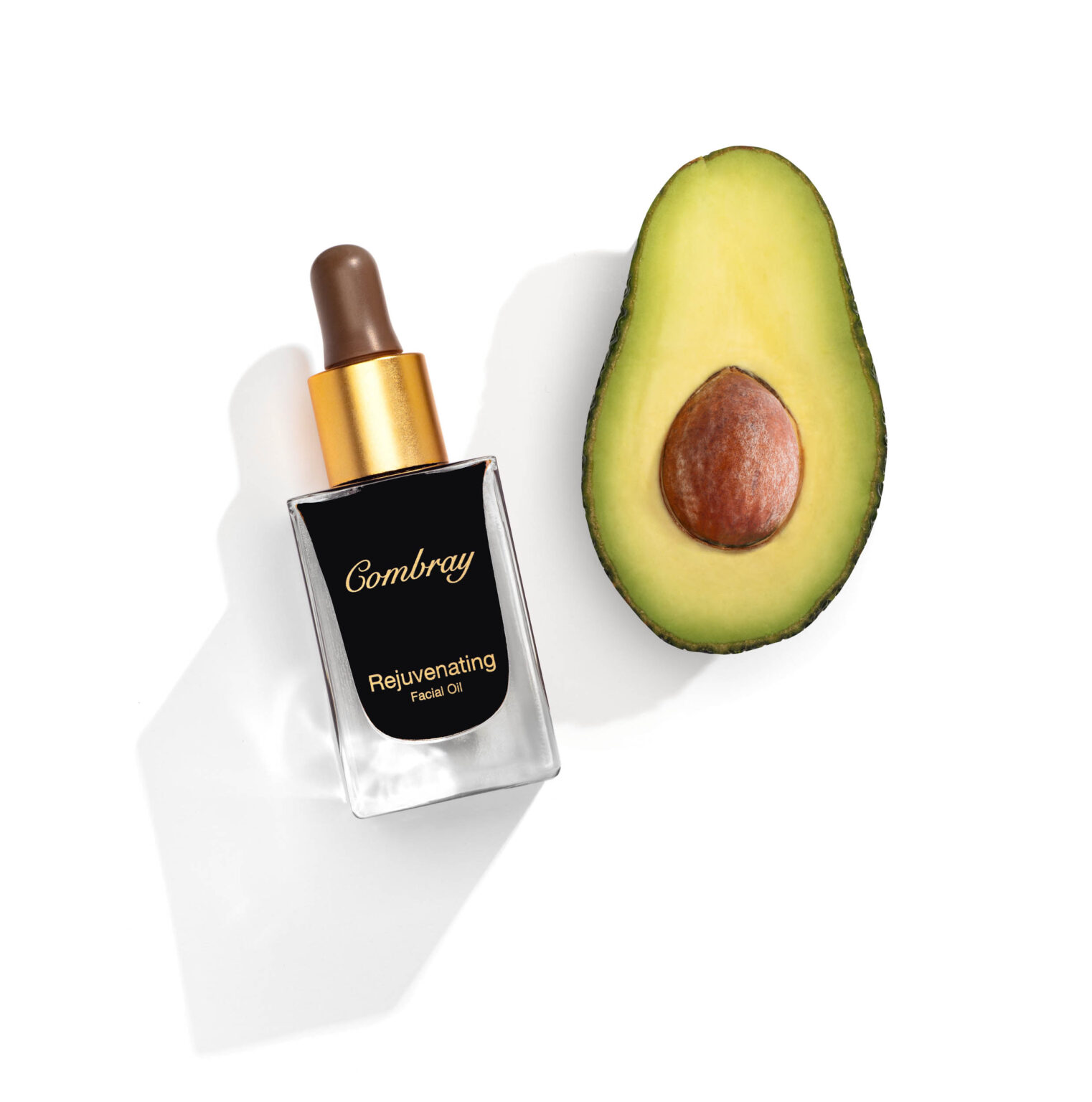 Een elegante compositie van een fles Combray Rejuvenating Facial Oil naast een halve avocado. De fles heeft een luxe uitstraling met goudkleurige accenten en een stijlvolle zwarte etiket. De avocado naast de fles voegt een natuurlijke en verfrissende touch toe aan de afbeelding. Fotografie door Jan Buwalda, Groningen.