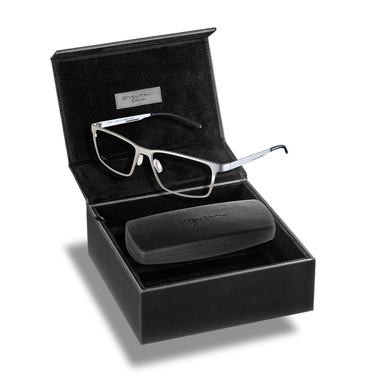Het beeld toont een stijlvolle bril verpakt in een elegante zwarte doos. De bril heeft een modern ontwerp met een slank, metalen montuur en subtiele accenten bij de pootjes. Naast de bril is er een bijpassende brillenkoker geplaatst, eveneens in elegant zwart uitgevoerd. De binnenkant van de doos is bekleed met fluweelachtig materiaal voor een luxe uitstraling. Fotografie door Jan Buwalda, Groningen.