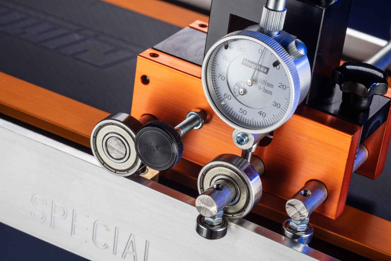 Dit is een close-upfoto van een precisie-instrument met een meetklok en metalen rolmechanismen. Het instrument is op een oranje aluminium basis bevestigd. Onderdelen zoals schroeven en lagers zijn duidelijk zichtbaar, en het metalen oppervlak is gemarkeerd met de tekst SPECIAL. De foto legt de technische aspecten van het apparaat vast met een gedetailleerde focus. Fotograaf: Jan Buwalda, Groningen.