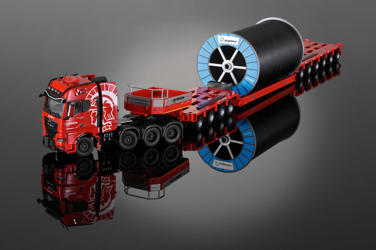 Deze foto toont een gedetailleerd schaalmodel van een rode vrachtauto met oplegger, die een grote zwarte kabelrol vervoert. De vrachtauto heeft opvallende witte versieringen en is voorzien van meerdere assen voor zware ladingen. De reflectie op de glanzende ondergrond versterkt het visuele effect. Fotograaf: Jan Buwalda, Groningen.