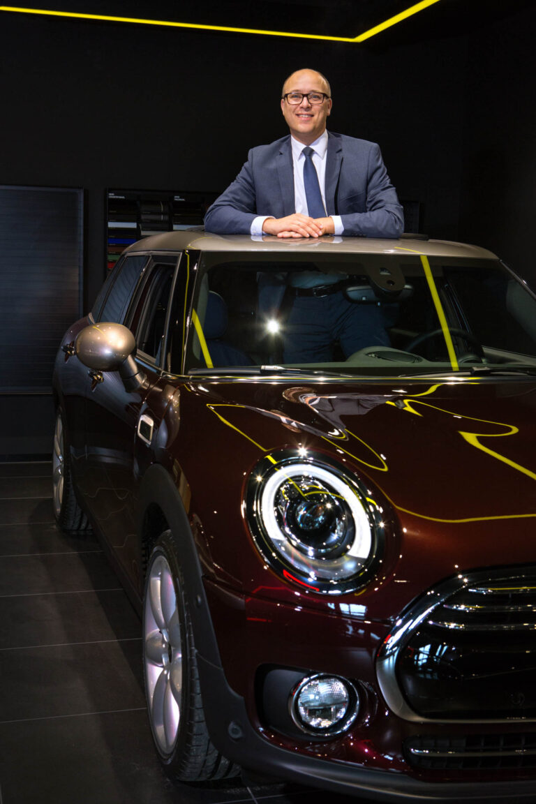Een man in een net pak leunt glimlachend uit het schuifdak van een elegante, donkerrode auto in een stijlvolle showroom. De auto heeft opvallende ronde koplampen en een glanzende afwerking. De sfeer is modern en professioneel, met strakke lijnen en subtiele verlichting. Fotograaf: Jan Buwalda, Groningen.