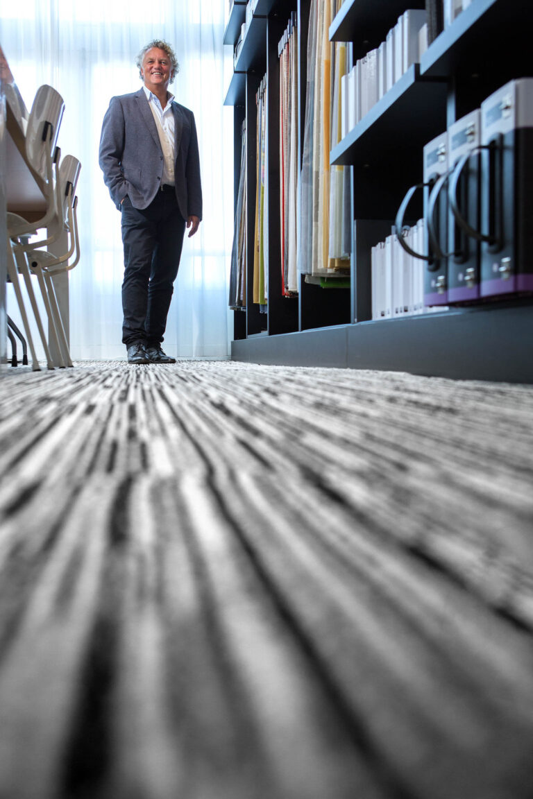 Dit is een foto van een man die in een moderne, goed verlichte ruimte staat met lange gangen en rekken vol met mappen en stoffen. Hij draagt een blauw colbert en zwarte broek, en kijkt glimlachend naar de camera. De vloer heeft een interessante textuur die in de compositie naar voren komt. De sfeer is professioneel en vriendelijk. Fotograaf: Jan Buwalda, Groningen.