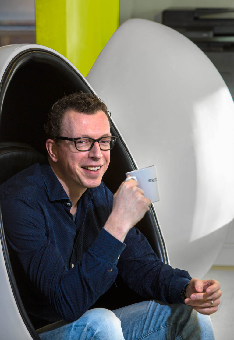 Het beeld toont een man die ontspannen zit in een moderne, eivormige stoel, terwijl hij een kopje vasthoudt. Hij draagt een donkerblauw overhemd en een bril, en glimlacht naar de camera. De achtergrond is neutraal met een opvallend geel element. Fotografie door Jan Buwalda, Groningen.