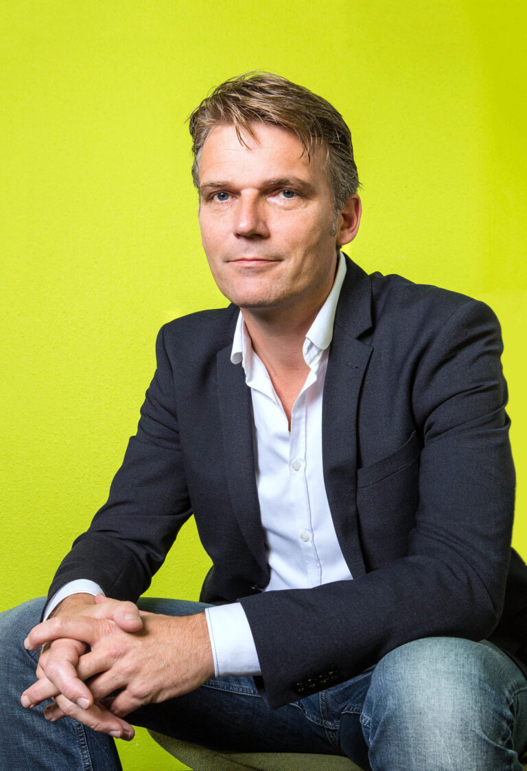 Het betreft een portretfoto van een man die voor een felgroene achtergrond zit. Hij draagt een donker jasje over een wit overhemd, met een nonchalante houding terwijl hij zijn handen in elkaar gevouwen heeft. De foto is helder verlicht, waardoor de kleuren van zijn kleding en de achtergrond sterk contrasteren. Fotograaf: Jan Buwalda, Groningen.