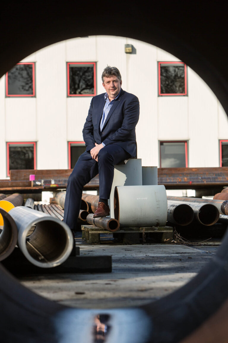 Dit is een foto van een man in een donkerblauw pak die ontspannen zit op een stapel industrieel materiaal, omgeven door grote buizen. Op de achtergrond is een gebouw met rode kozijnen zichtbaar. De foto is gemaakt door Jan Buwalda in Groningen.