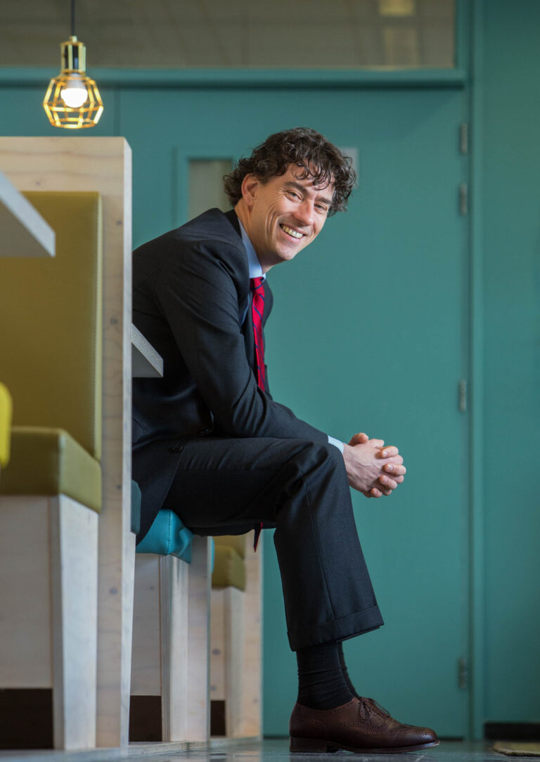 Ik ben sorry, ik weet niet wie deze persoon is.Beschrijving: Een man in een donkergrijs pak zit ontspannen op een moderne, houten bank. Hij lacht vriendelijk terwijl hij naar de camera kijkt. De achtergrond is een muur in een frisse, blauwe tint. Een stijlvolle lamp hangt boven de ruimte waar hij zit. De foto straalt een professionele en toegankelijke sfeer uit. Fotograaf: Jan Buwalda, Groningen.