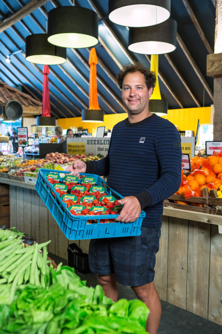 In een groentewinkel in Groningen staat een glimlachende man met een blauw gestreepte trui en korte broek. Hij houdt een blauwe krat vast met daarin meerdere bakjes aardbeien van het merk Melkard. Op de achtergrond zijn verscheidene fruit- en groentensoorten te zien met felgekleurde lampen die boven de houten toonbank hangen. De sfeer is kleurrijk en uitnodigend. De foto is gemaakt door Jan Buwalda.