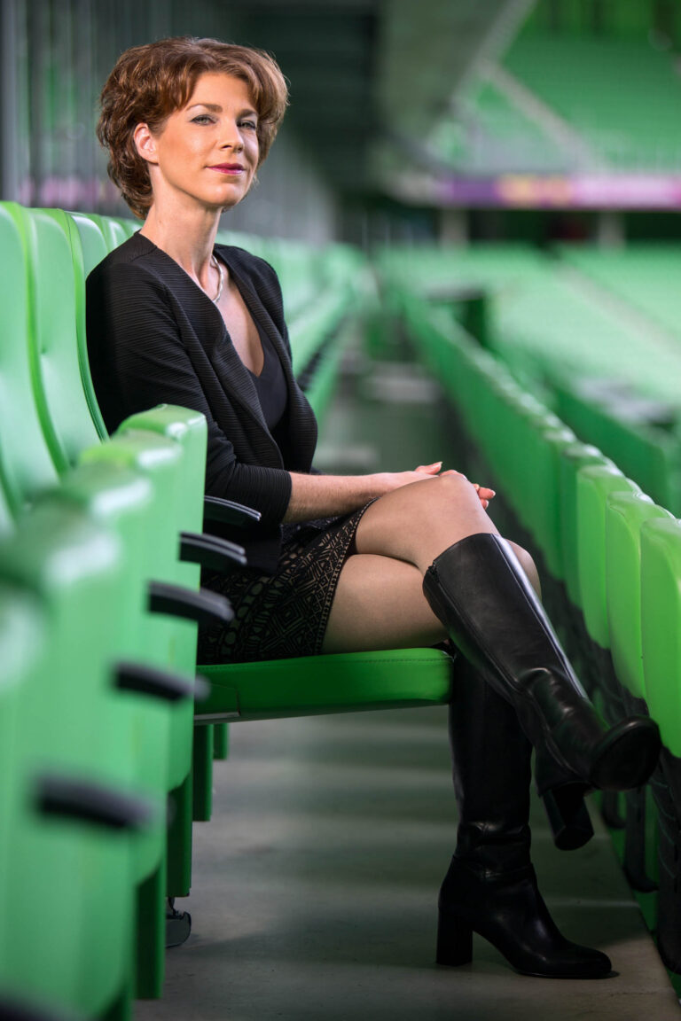 Het is een portretfoto van een vrouw die op een groene stadionstoel zit. Ze draagt een zwarte outfit met een patroonrok en hoge zwarte laarzen. De achtergrond vervaagt verder in een stadionomgeving, waardoor de groene stoelen in contrast staan met haar kleding. De foto is gemaakt door Jan Buwalda in Groningen.
