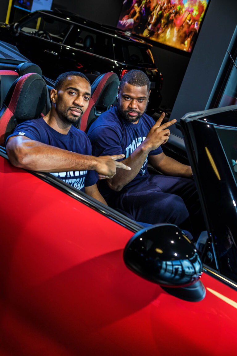 Twee mannen zitten in een sportieve rode auto. Ze dragen beiden donkerblauwe T-shirts en maken peace-tekens met hun handen. De auto bevindt zich in een moderne omgeving, mogelijk een showroom, met een donkere auto op de achtergrond en een kleurrijk kunstwerk aan de muur. Foto door Jan Buwalda, Groningen.