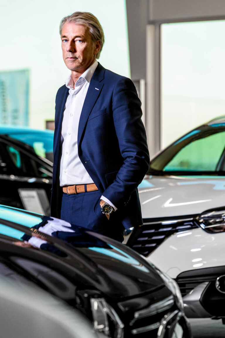Dit is een zakelijke portretfoto van een man in een showroom vol auto's. De man draagt een stijlvol donkerblauw pak met een witte overhemd en een bruine riem. Hij heeft een horloge om zijn pols en kijkt serieus richting de camera. Op de achtergrond zijn verschillende auto's te zien onder de heldere verlichting van de showroom. Deze foto is genomen door fotograaf Jan Buwalda in Groningen.