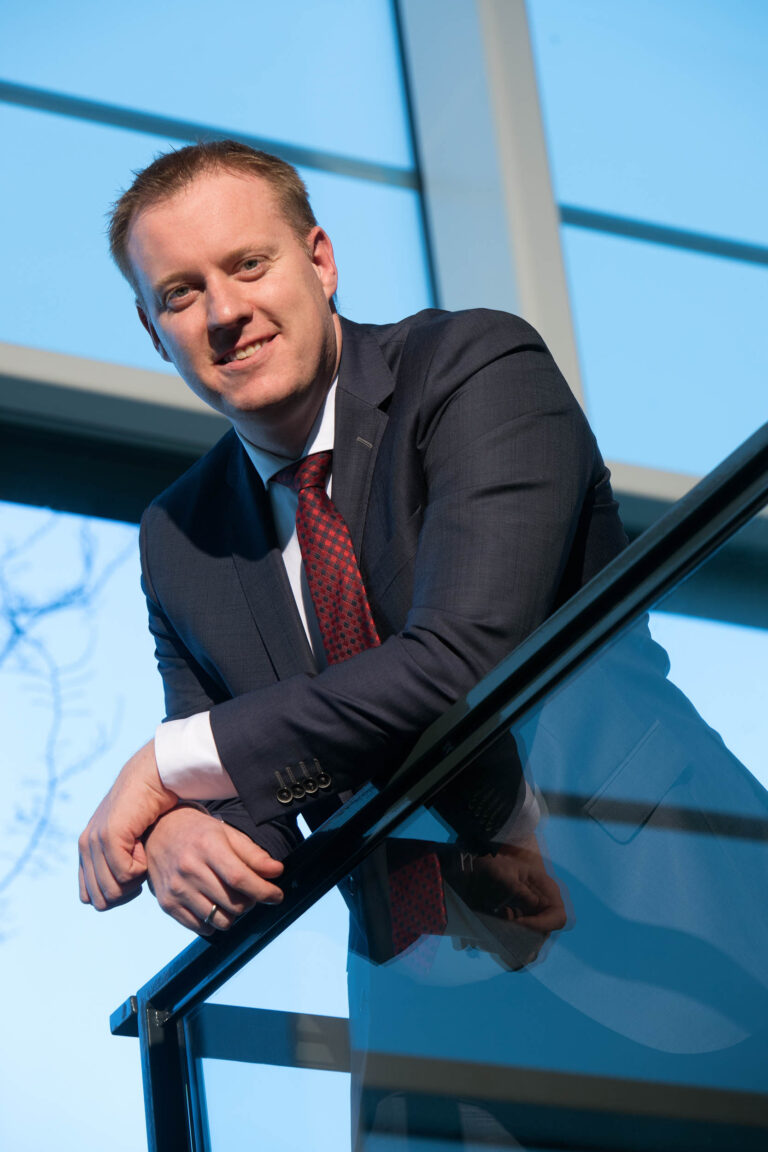 Het beeld toont een man gekleed in een donkerblauw pak met een rood geruite stropdas, die leunt op een moderne glazen balustrade. De achtergrond is helderblauw met grote ramen die buitenlicht binnenlaten. De persoon heeft een vriendelijke glimlach en de foto straalt een professionele sfeer uit. Fotograaf: Jan Buwalda, Groningen.