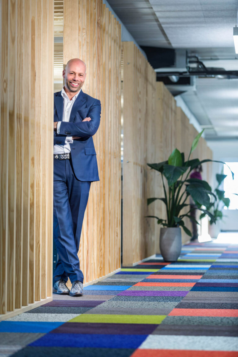 Dit portret toont een man in een blauw pak die ontspannen leunt tegen een houten wand in een moderne kantooromgeving. De kleurrijke, geometrische vloerbedekking voegt een speelse sfeer toe, terwijl de grote plant aan de rechterkant een natuurlijke touch biedt. De foto is gemaakt door Jan Buwalda in Groningen.