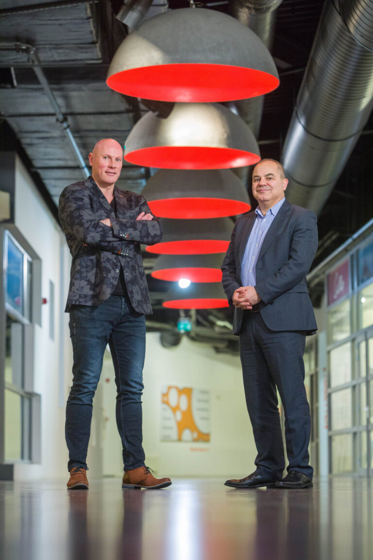 Dit is een foto gemaakt door Jan Buwalda in Groningen. Twee mannen staan in een moderne gang. De ene man draagt een donker colbert met een patroon, jeans en bruine schoenen, en staat met gekruiste armen. De andere man heeft een donker colbert en pantalon aan met een lichtblauw overhemd. Boven hen hangen grote lampen met rode binnenkanten. De ruimte heeft een industriële uitstraling met zichtbare leidingen aan het plafond.