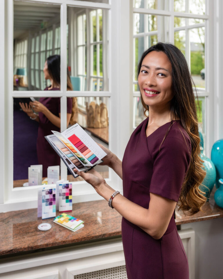 Het beeld toont een stijlvolle vrouw in een paarse jurk, glimlachend terwijl ze staande bij een raam naar een kleurenwaaier kijkt. Ze bevindt zich in een ruimte met veel natuurlijk licht, en haar reflectie is te zien in de grote ramen. Op de achtergrond zijn er folders en kleurkaarten op een tafelblad zichtbaar. Fotograaf: Jan Buwalda, Groningen.