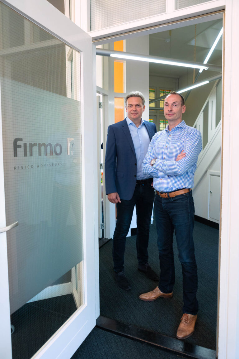 Het beeld toont twee mannen die voor de ingang van een kantoorruimte staan. De linkerman draagt een net donkerblauw pak met een lichtblauw overhemd eronder, terwijl de rechterman in een geruite blauwe blouse en donkere jeans gekleed is. Het kantoor heeft een deur met het logo van Firmo Risico Adviseurs erop. De omgeving is modern met lichte kleuren en een nette uitstraling. De foto is genomen door Jan Buwalda in Groningen.