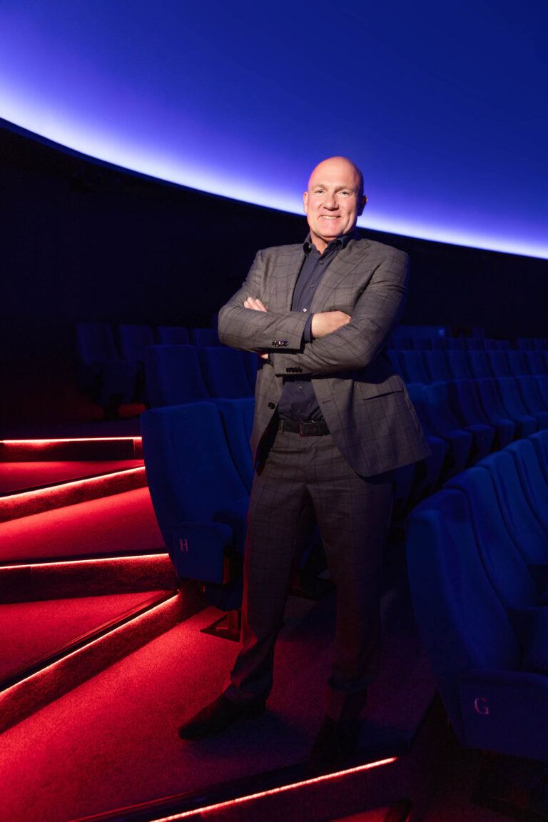 In deze foto staat een man in een formeel grijs pak met gekruiste armen. Hij staat in een moderne bioscoopzaal met een opvallend verlichte, halfronde blauwe koepel op de achtergrond. De vloer is verlicht met rode lichten die de rijen blauwe stoelen accentueren. De sfeer is professioneel en futuristisch. Fotograaf: Jan Buwalda. Plaats: Groningen.