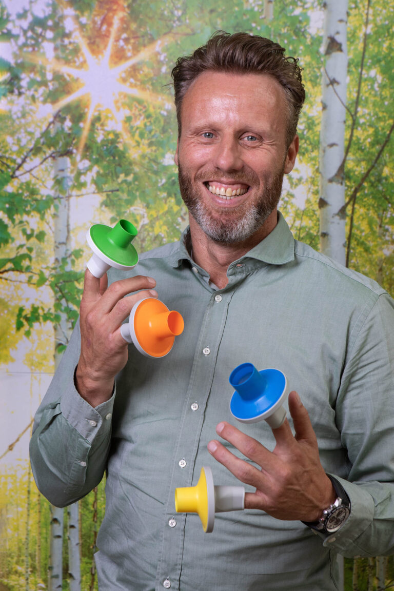 Dit portret toont een glimlachende man in een groene overhemden die vier kleurrijke objecten in zijn handen houdt. De achtergrond is een levendige afbeelding van een bos met zonlicht dat door de bladeren schijnt. Fotografie door Jan Buwalda in Groningen.