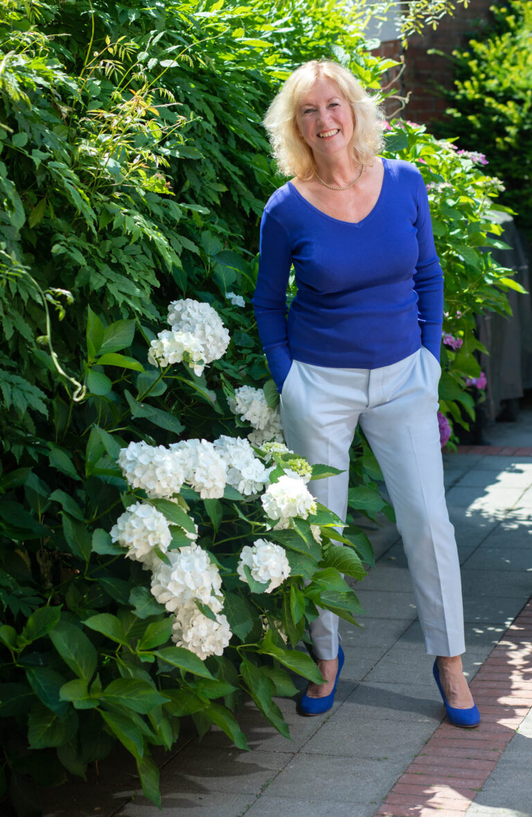 Dit is een kleurrijke foto van een vrouw die ontspannen poseert in een weelderige tuin. Ze draagt een blauwe top en witte broek, terwijl ze staat naast een struik met witte bloemen. De achtergrond is rijkelijk gevuld met groene bladeren, wat een levendige en frisse sfeer creëert. De compositie straalt een gevoel van rust en natuurlijke schoonheid uit. Fotograaf: Jan Buwalda, Groningen.