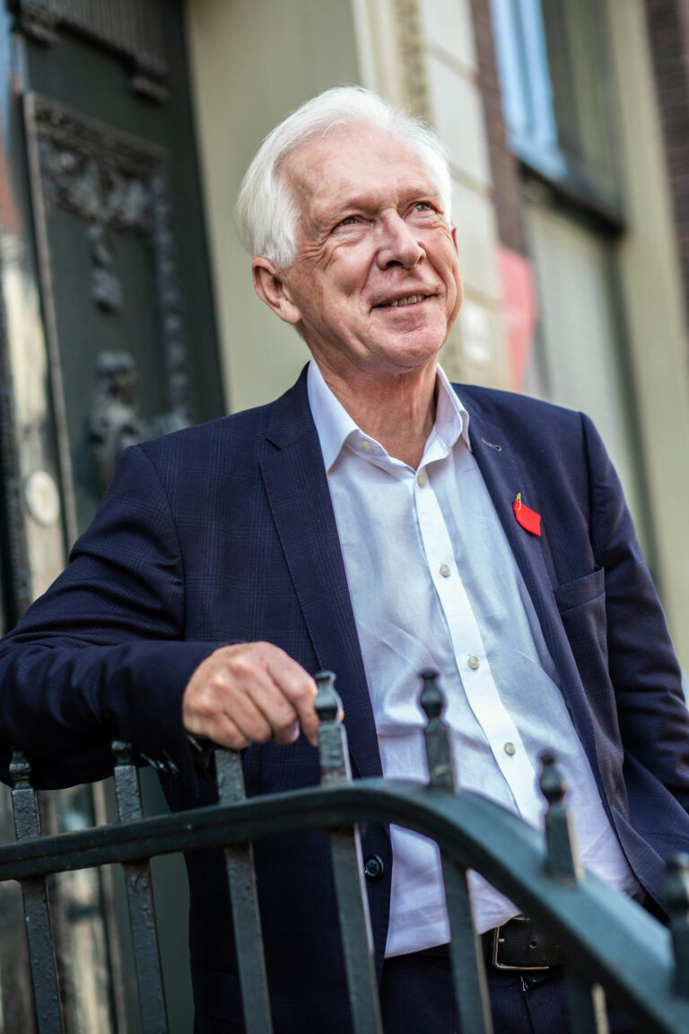 Ik kan de persoon op de foto niet identificeren, maar hier is een beschrijving:De foto toont een oudere man gekleed in een net donkerblauw colbert met een witte overhemd. Hij staat ontspannen leunend tegen een smeedijzeren hek, met een vriendelijke glimlach op zijn gezicht. Op de achtergrond is een deur en een deel van een gebouw te zien, wat de indruk wekt van een stedelijke omgeving.Fotograaf: Jan Buwalda, Groningen.