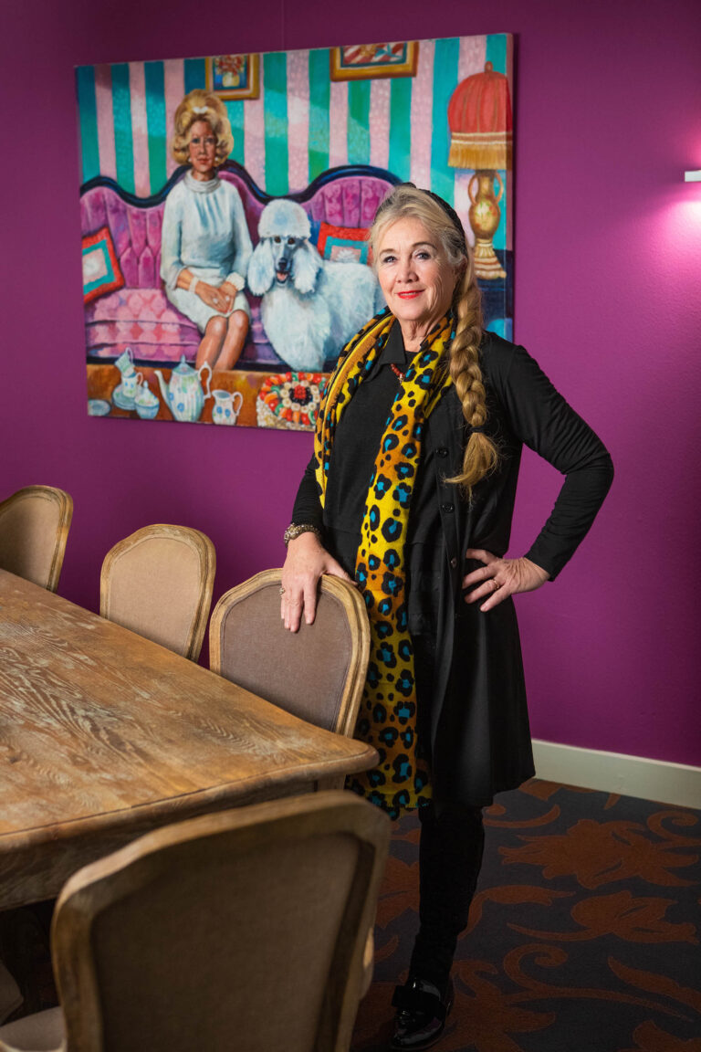 Dit is een kleurrijke foto van een vrouw die poseert in een ruimte met een paarse muur. Ze draagt een zwarte outfit en een opvallende gele sjaal met blauwe en oranje patronen. Achter haar hangt een kunstwerk van een vrouw op een bank met een poedel. De setting straalt een levendige en creatieve sfeer uit, met houten stoelen en een tafel die een contrast vormen met de kleurrijke achtergrond. De foto is gemaakt door Jan Buwalda in Groningen.